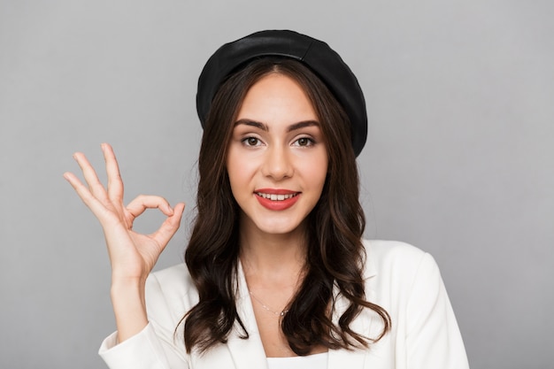 Portrait d'une belle jeune femme portant béret debout isolé sur fond gris, montrant le geste ok