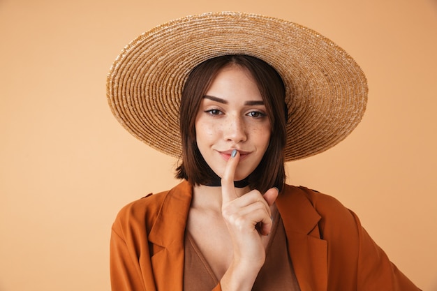 Portrait d'une belle jeune femme portant un chapeau de paille debout isolé sur un mur beige, montrant un geste de silence