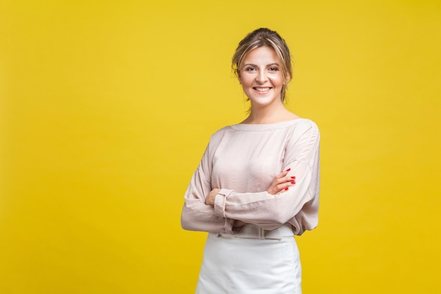 Portrait d'une belle jeune femme positive aux cheveux blonds en chemisier beige décontracté debout avec les bras croisés et regardant la caméra, sourire à pleines dents. studio d'intérieur tourné isolé sur fond jaune