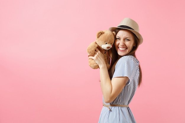 Portrait de belle jeune femme en robe bleue, chapeau de paille d'été tenant un ours en peluche isolé sur fond rose. Personnes, émotions sincères, concept de style de vie. Espace publicitaire. Espace de copie.