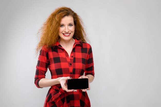 Portrait de belle jeune femme rousse en robe à carreaux souriant montrant téléphone mobile