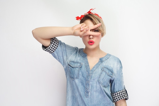 Portrait d'une belle jeune femme sexuelle en chemise en jean bleu décontractée avec maquillage et bandeau rouge debout, montrant la victoire v chanter avec le doigt. tourné en studio intérieur, isolé sur fond blanc