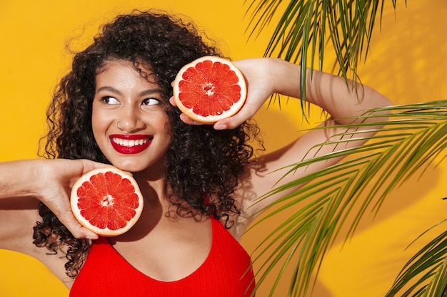 Portrait d'une belle jeune femme souriante en forme portant un bikini debout isolée sur un mur exotique jaune, posant avec un pamplemousse coupé en deux