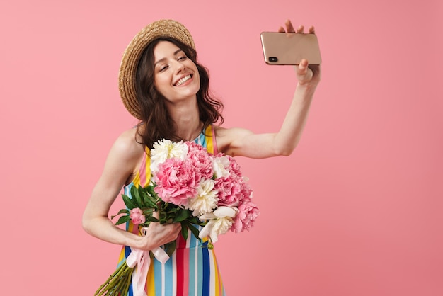 Portrait d'une belle jeune femme souriante et souriante posant isolée sur un mur rose tenant des fleurs prendre un selfie par téléphone portable