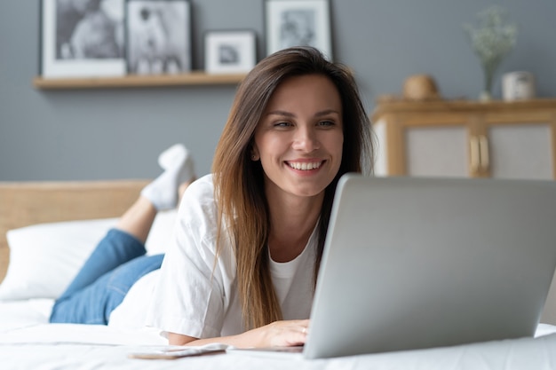 Portrait d'une belle jeune femme souriante utilisant un ordinateur portable au lit à la maison.