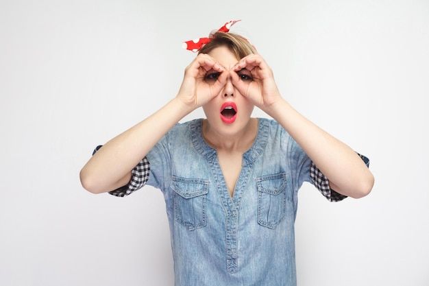 Portrait d'une belle jeune femme surprise en chemise en jean bleue décontractée avec maquillage et bandeau rouge debout avec des jumelles geste des mains et à la recherche. tourné en studio intérieur, isolé sur fond blanc