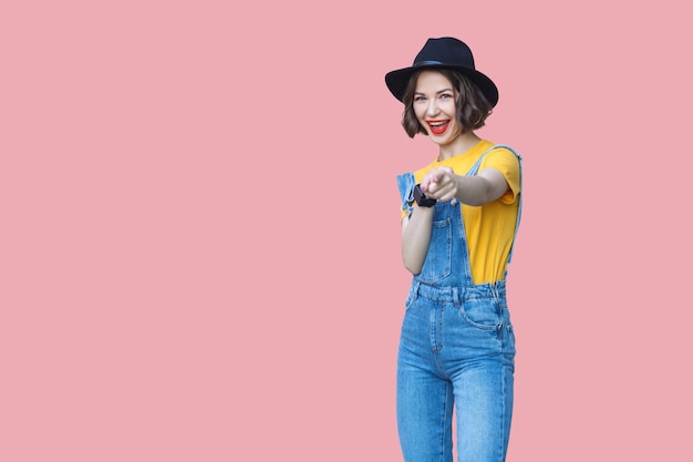 Portrait d'une belle jeune femme surprise en t-shirt jaune, salopette en jean bleu avec maquillage et chapeau noir debout, étonné, pointant et regardant la caméra. tourné en studio isolé sur fond rose.