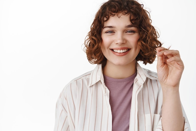 Portrait de la belle jeune femme touchant les cheveux courts bouclés et souriant, fait une nouvelle coupe de cheveux au salon de beauté du coiffeur, debout heureux sur fond blanc.