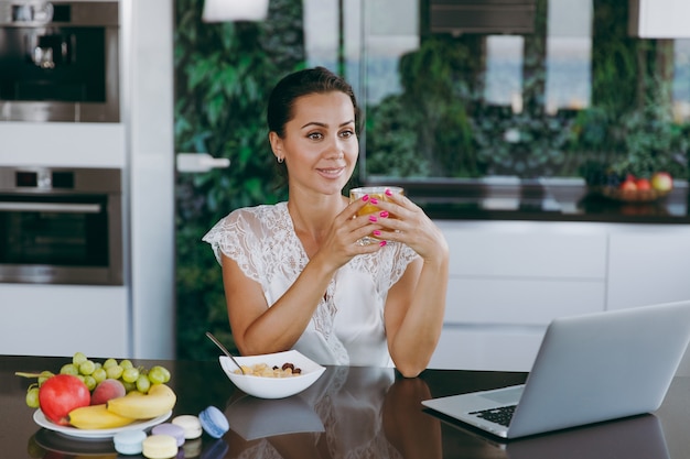 Le portrait d'une belle jeune femme travaillant avec un ordinateur portable pendant le petit-déjeuner avec des céréales et du lait et buvant du jus d'orange