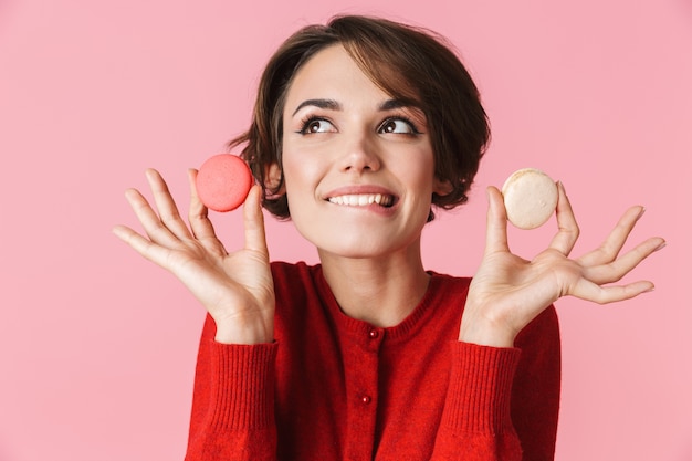 Portrait d'une belle jeune femme vêtue d'une robe rouge debout isolé sur fond rose, tenant des macarons