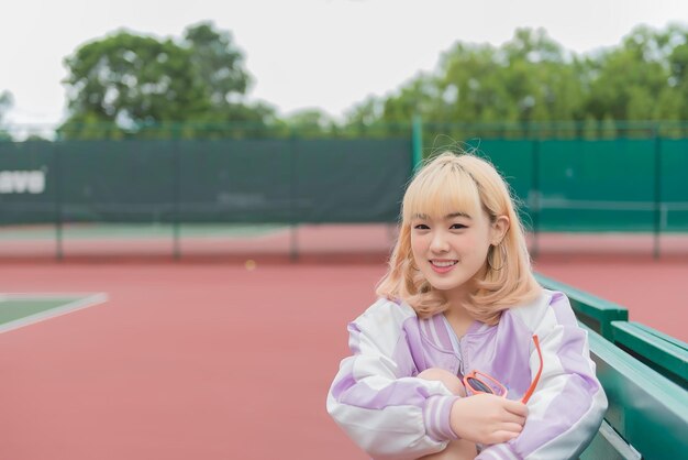 Portrait d'une belle jeune fille aux cheveux dorés assis sur le banc du court de tennis Jolie femme pose pour prendre une photo à l'extérieur style kawaii