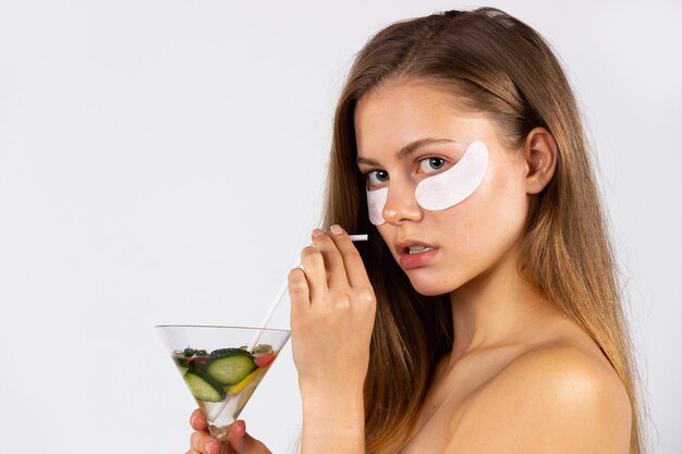 Portrait d'une belle jeune fille aux cheveux longs sous les yeux avec des pansements oculaires et un cocktail dans ses mains. Soins de la peau et concept d'aliments sains. Photo sur mur blanc. Photo de haute qualité