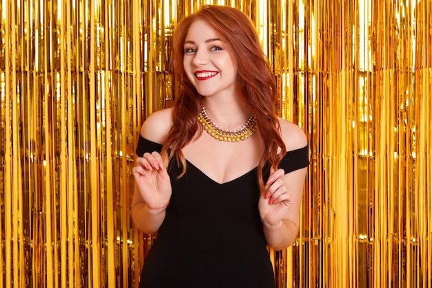 Portrait de la belle jeune fille aux cheveux rouges en robe noire posant contre le mur décoré de guirlandes de paillettes, regardant la caméra avec le sourire aux lèvres rouges.