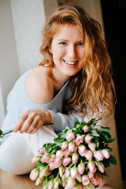 Portrait d'une belle jeune fille avec un bouquet de roses près de la fenêtre. Cheveux roux bouclés.