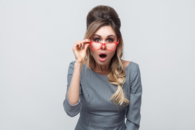 Portrait d'une belle jeune fille choquée en robe grise avec une queue de cochon sur la tête debout, portant des lunettes rouges et regardant la caméra avec un visage étonné. Intérieur, isolé, tourné en studio, fond gris