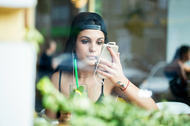 Portrait d'une belle jeune fille. Elle est assise seule dans un café, écoute de la musique au casque et vérifie le maquillage à travers l'appareil photo d'un smartphone. Tiré à travers la fenêtre.
