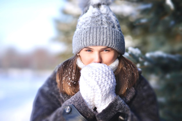 Portrait de belle jolie fille jeune jolie femme congelée debout marchant dans le parc enneigé d'hiver à la neige froide journée glaciale en chapeau, écharpe, pull blanc se réchauffant les mains dans des gants en regardant la caméra