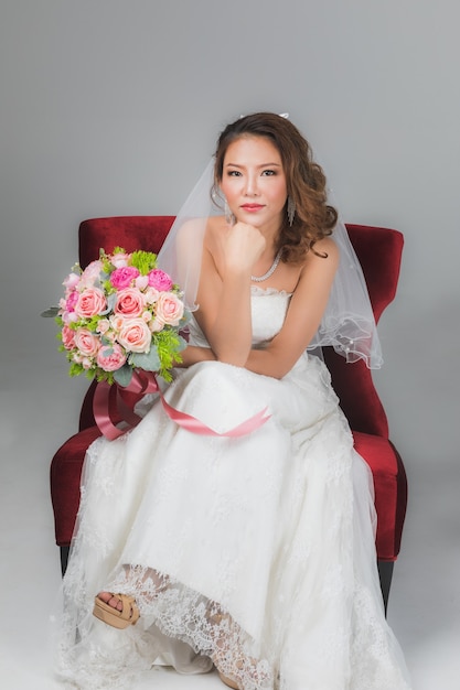 Portrait d'une belle mariée asiatique assise sur une chaise rouge tenant un bouquet de fleurs et mettant sa main sous le menton sur fond noir gris.