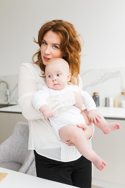 Portrait d'une belle mère debout et regardant joyeusement à huis clos tout en tenant son mignon petit bébé dans les mains