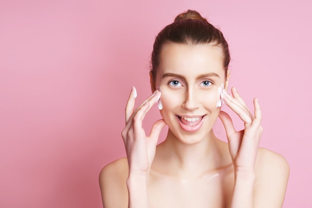 Portrait de belle modèle dame avec maquillage naturel, appliquer la crème sur son visage. isolé sur rose