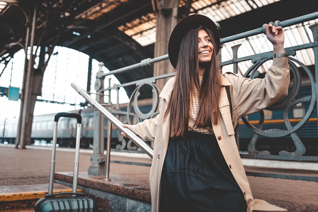 Portrait d'une belle passagère élégante positive en manteau et chapeau à la gare