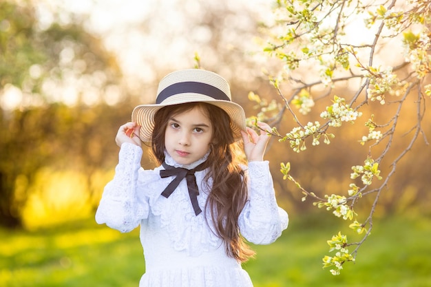 Portrait de la belle petite fille au chapeau et robe blanche debout sous les arbres en fleurs au printemps