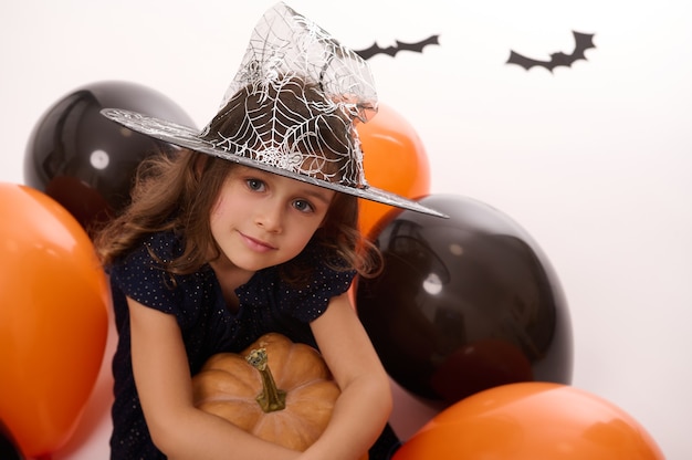 Portrait d'une belle petite fille portant un chapeau de sorcier, vêtue d'un costume de carnaval de sorcière sombre, embrasse une citrouille dans sa main, s'assoit sur un fond blanc avec des ballons et des chauves-souris orange noirs colorés