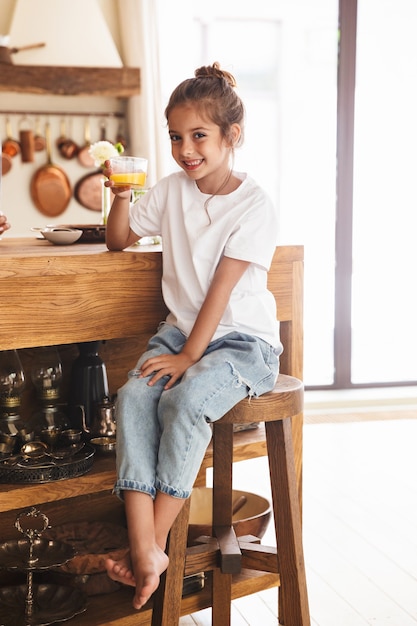 Portrait d'une belle petite fille prenant son petit déjeuner et buvant du jus d'orange assis à table dans une cuisine lumineuse à la maison