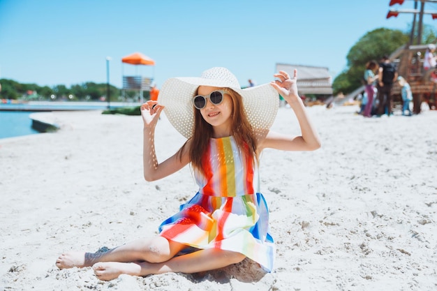 Portrait d'une belle petite fille vêtue d'une robe rayée et d'un chapeau de paille assis sur la plage