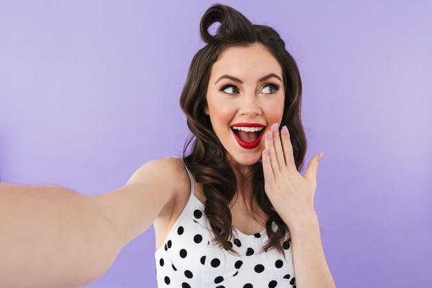 Portrait d'une belle pin-up portant un maquillage lumineux debout isolé sur un mur violet, prenant un selfie