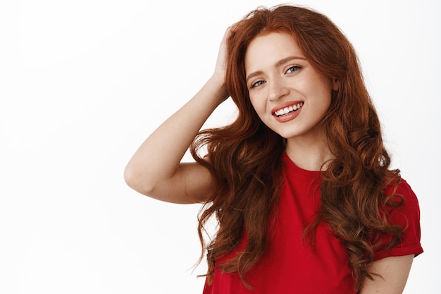 Portrait d'une belle rousse naturelle aux cheveux bouclés, touchant la tête et souriante heureuse. Femme candide avec un sourire blanc, portant un t-shirt rouge, debout sur fond blanc