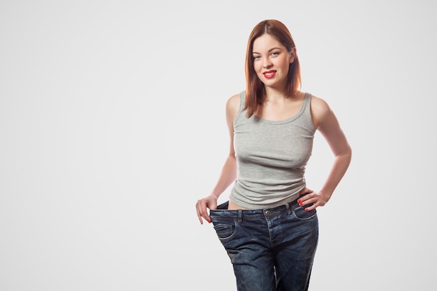 Portrait d'une belle taille mince heureuse de jeune femme en gros jeans et haut gris montrant une perte de poids réussie, intérieur, tourné en studio, isolé sur fond gris clair, concept de régime.