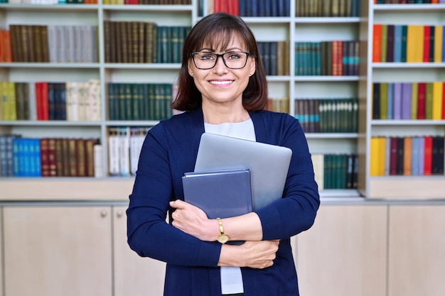 Portrait d'une bibliothécaire avec un ordinateur portable dans les mains dans la bibliothèque Une enseignante d'âge moyen souriante en lunettes regardant la caméra Professeur mentor tuteur posant dans le bureau de la classe