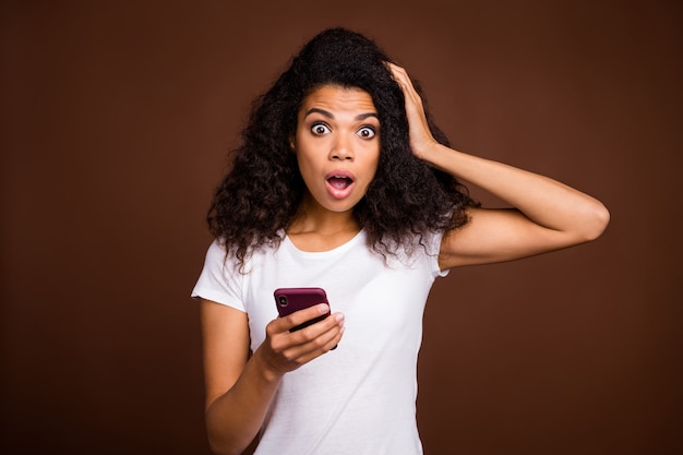 Portrait de blogueur fille afro-américaine étonné utiliser téléphone portable lire les informations stare stupeur main toucher la tête crier omg porter une tenue de style décontracté.
