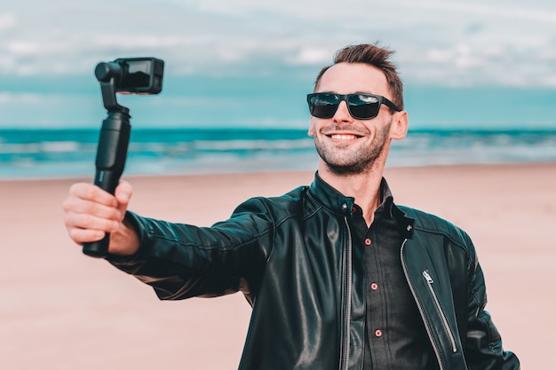 Portrait De Blogueur Souriant Dans Des Lunettes De Soleil Faisant Selfie Ou Vidéo En Streaming à La Plage à L'aide D'une Caméra D'action Avec Stabilisateur De Caméra à Cardan.