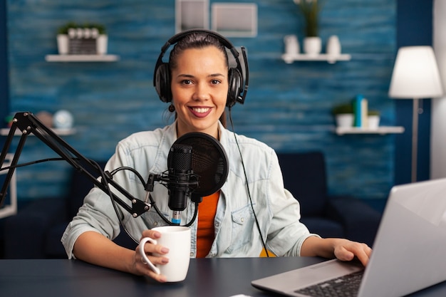 Portrait d'un blogueur souriant regardant la caméra avant de commencer une vidéo en direct sur un podcast home studio tenant une tasse de café. Une blogueuse créatrice de contenu enregistrant une diffusion en direct de brodcast pour Internet