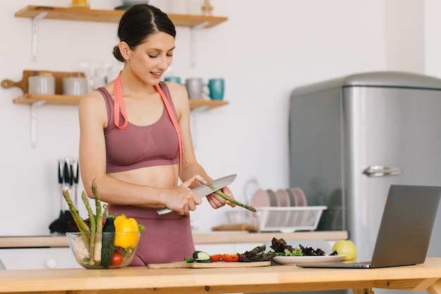 Portrait d'une blogueuse sportive heureuse faisant la cuisine des asperges et menant une vidéoconférence sur le sujet de l'alimentation saine sur un ordinateur portable dans la cuisine