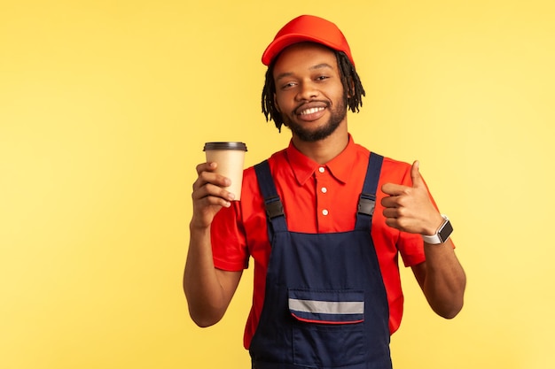 Portrait d'un bricoleur souriant vêtu d'un t-shirt rouge et d'une salopette bleue tenant du café pour aller dans les mains et montrant les pouces vers le haut en regardant la caméra Prise de vue en studio intérieur isolée sur fond jaune