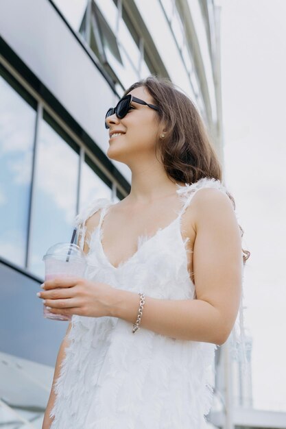 Portrait d'une brune qui boit des smoothies à l'extérieur tenue élégante robe blanche lunettes de soleil Ambiance estivale