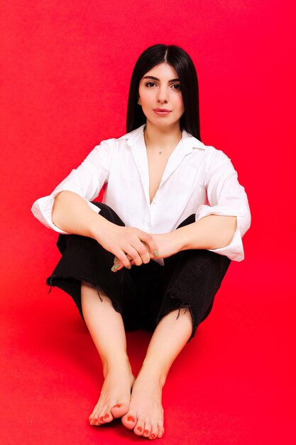 Portrait d'une brune vêtue d'une chemise blanche et d'un jean foncé assise sur un fond rouge Studio de photographie de mode