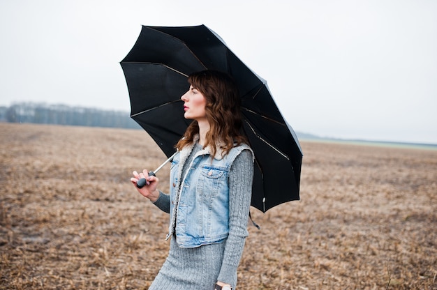 Portrait de brunette fille bouclée en veste de jeans avec un parapluie noir au champ.