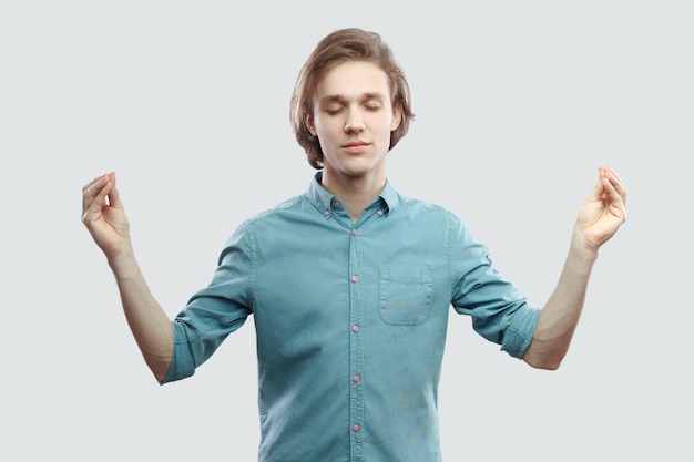 Portrait de calme beau jeune homme blond aux cheveux longs en chemise décontractée bleue debout dans une pose de yoga et méditant, et essayez de vous détendre. tourné en studio intérieur, isolé sur fond gris clair.