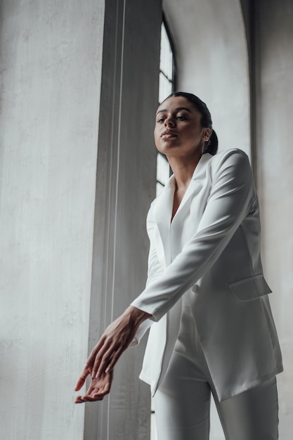 Portrait de candide authentique fille modèle métisse sexy en tailleur-pantalon blanc posant la main sur le visage