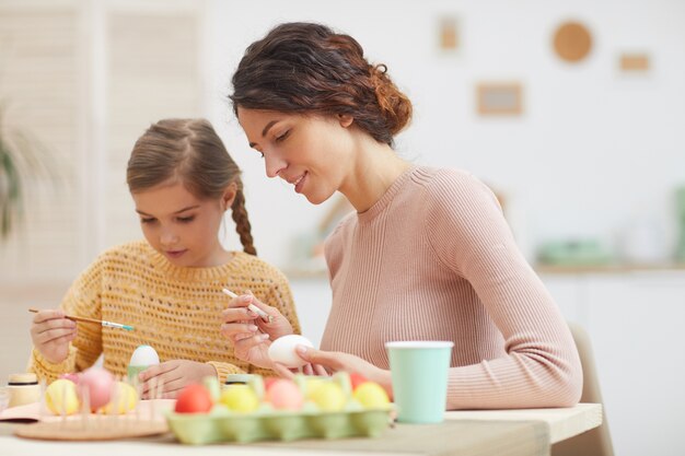 Portrait candide de la mère et la fille peinture des oeufs de Pâques assis à table dans l'intérieur de la cuisine confortable, espace copie