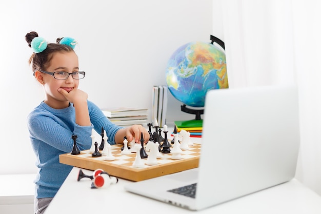 Portrait Caucasian Petite fille cirly jouant aux échecs à la table à l'intérieur avec échiquier. petite fille apprend les échecs sur un ordinateur portable en ligne, apprentissage à distance