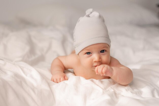 Portrait d'un charmant bébé joyeux aux yeux bleus dans un bonnet blanc allongé sur le ventre sur du linge de lit blanc. espace pour le texte. Photo de haute qualité