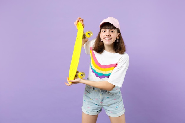 Portrait d'une charmante adolescente souriante dans des vêtements vifs tenant une planche à roulettes jaune isolée sur fond de mur pastel violet en studio. Émotions sincères des gens, concept de style de vie. Maquette de l'espace de copie.