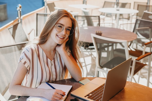 Portrait d'une charmante femme aux cheveux rouges et taches de rousseur souriant tout en travaillant en plein air à son ordinateur portable dans un café.