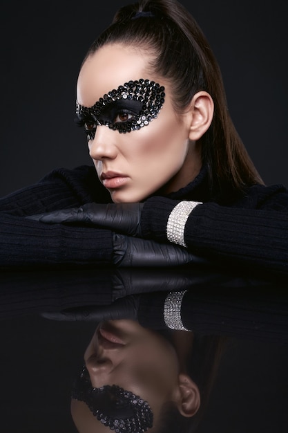 Portrait de charmante femme brune élégante en pull à col roulé noir et masque de paillettes posant sur une surface en miroir en studio