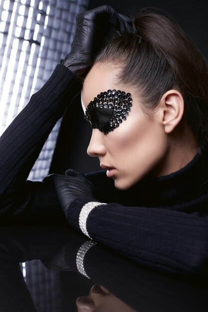 Portrait de charmante femme brune élégante en pull à col roulé noir et masque de paillettes posant sur une surface en miroir en studio